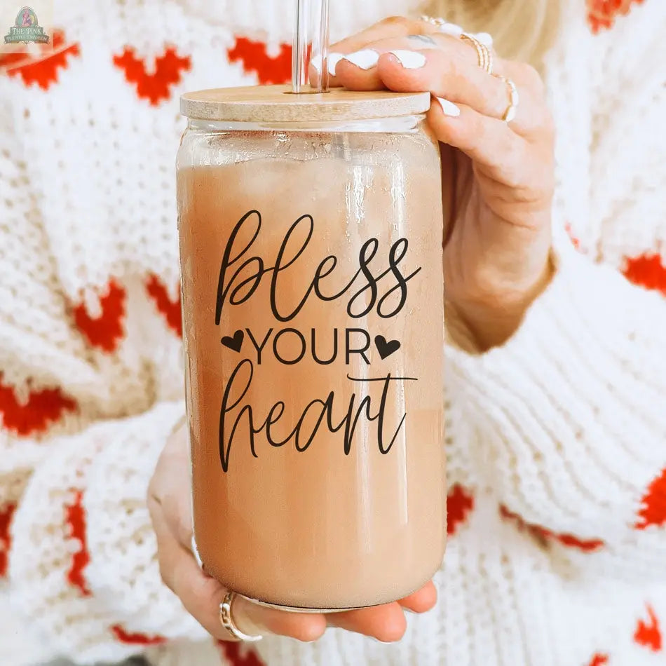 A person in a white sweater with red hearts holds a light brown drink in the Bless Your Heart 20oz glass cup, which has a bamboo lid, straw, and the phrase "bless your heart" on it.