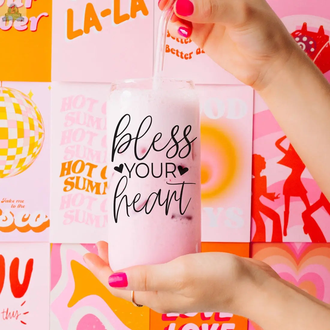 A person with pink nail polish holds the Bless Your Heart 20oz glass cup, filled with a pink iced drink and a straw. Retro posters in orange, pink, and red hues decorate the background.