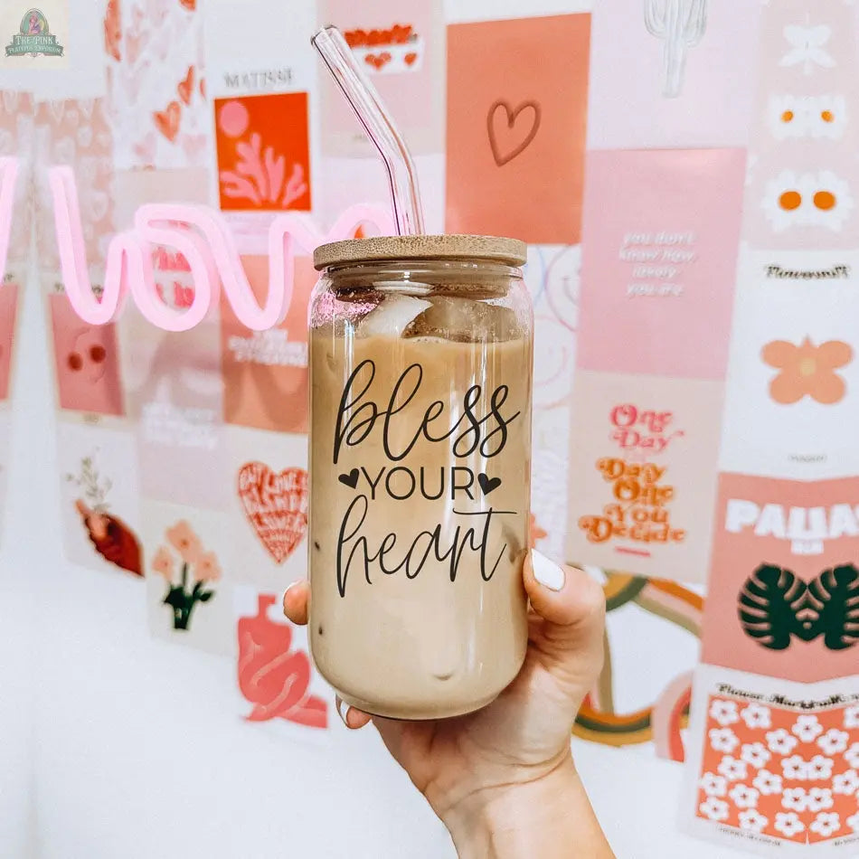 A hand holds the Bless Your Heart 20oz glass cup filled with iced coffee, featuring a bamboo lid and straw. In the background, there’s a pink-themed art print collage and a neon love sign decorating the wall.