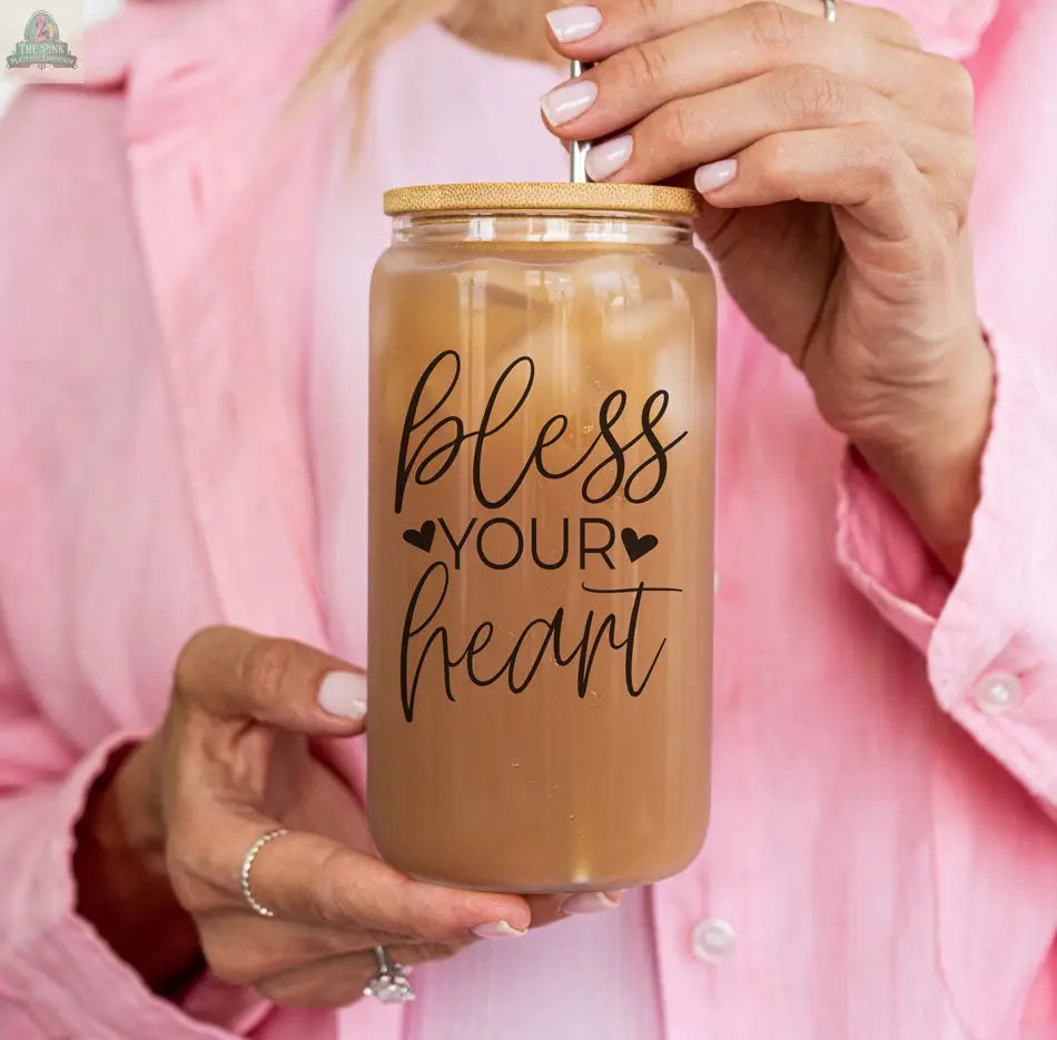 Someone in a light pink shirt holds the Bless Your Heart 20oz glass cup, filled with iced coffee and a metal straw. This cup features small heart designs, adding a charming touch to any drinkware collection.