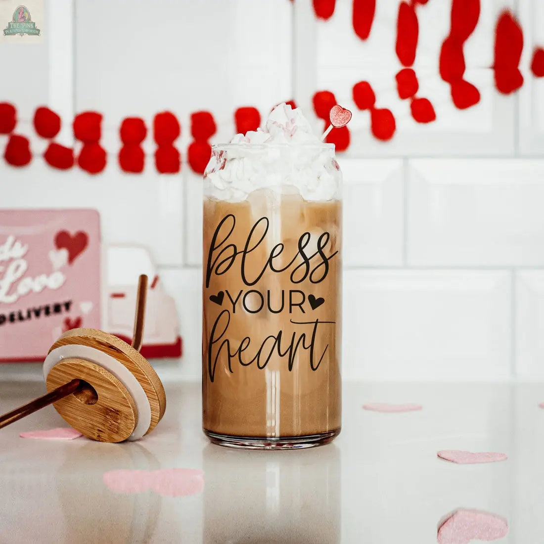 A Bless Your Heart 20oz glass cup brimming with iced coffee and whipped cream sits on a counter, surrounded by red and pink Valentine’s hearts—a festive bamboo lid glass cup perfect for the season.