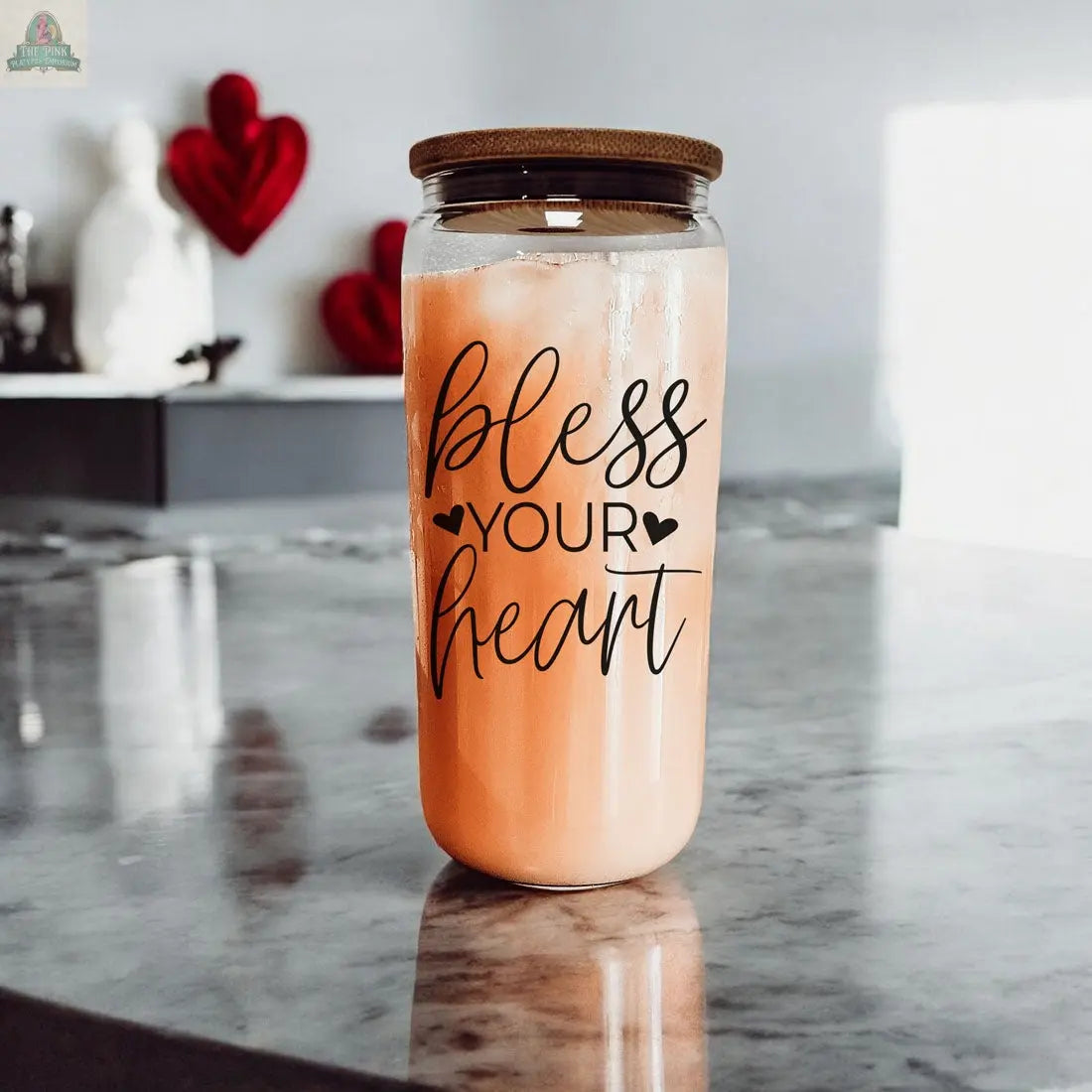 The Bless Your Heart 20oz glass cup with a bamboo lid holds an iced pink drink on a marble counter. The jar features black script and two heart icons, with blurred red heart decorations in the background.