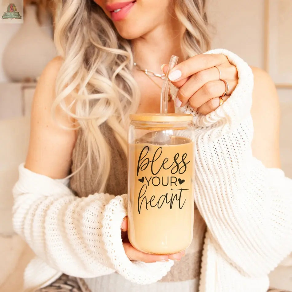 A woman with long blonde hair and a cozy white cardigan smiles softly while holding a Bless Your Heart 20oz glass cup filled with iced coffee. She has nude nail polish and enjoys her drink.