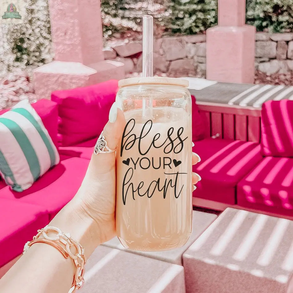 A hand holds the Bless Your Heart 20oz glass cup with a straw. Behind it, vibrant pink outdoor couches with striped cushions are lit by sunlight, creating playful shadows.