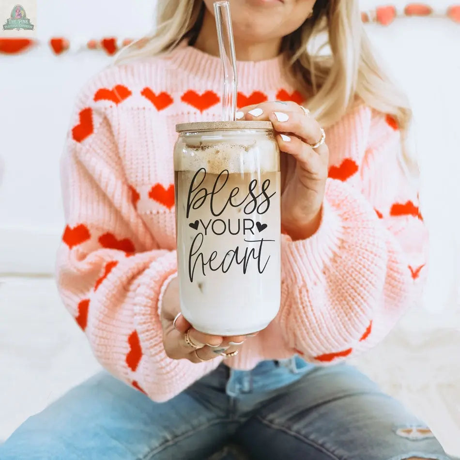 A woman in a pink sweater with red hearts sits cross-legged on a bed, holding a Bless Your Heart 20oz glass cup filled with iced coffee and a straw.