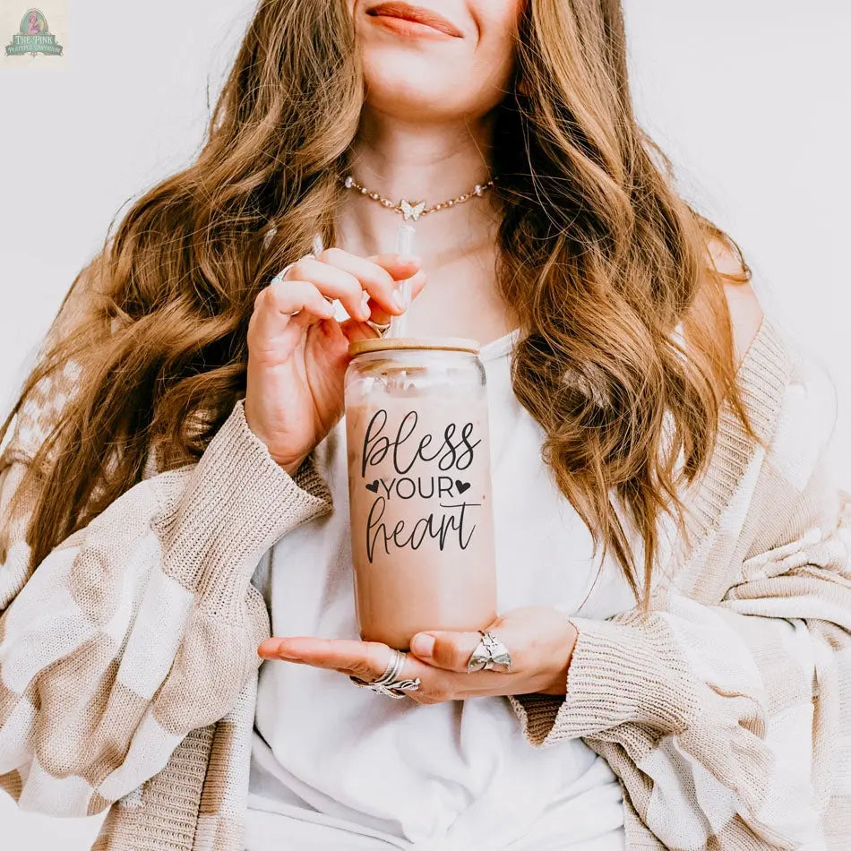 A woman with long wavy hair, wearing a cream cardigan and several rings, smiles softly while holding a Bless Your Heart 20oz glass cup of iced coffee with a bamboo lid and straw.