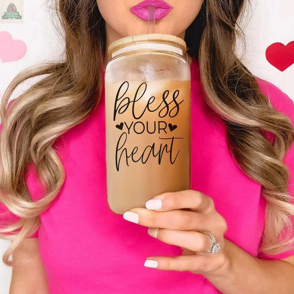 A woman in a pink shirt drinks from a Bless Your Heart 20oz glass cup with a bamboo lid and pink straw, with pink and red heart decorations in the background.