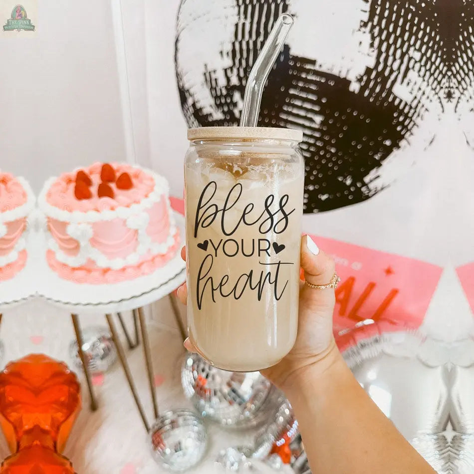 A hand holds the Bless Your Heart 20oz glass cup with a clear straw and bamboo lid, surrounded by pink heart-shaped cakes, disco balls, and festive decorations for a cheerful atmosphere.