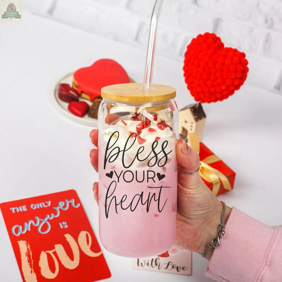 A hand holds the Bless Your Heart 20oz glass filled with a pink milkshake topped with whipped cream, heart sprinkles, and a straw. Red heart decorations, a card, and small gifts are in the background.