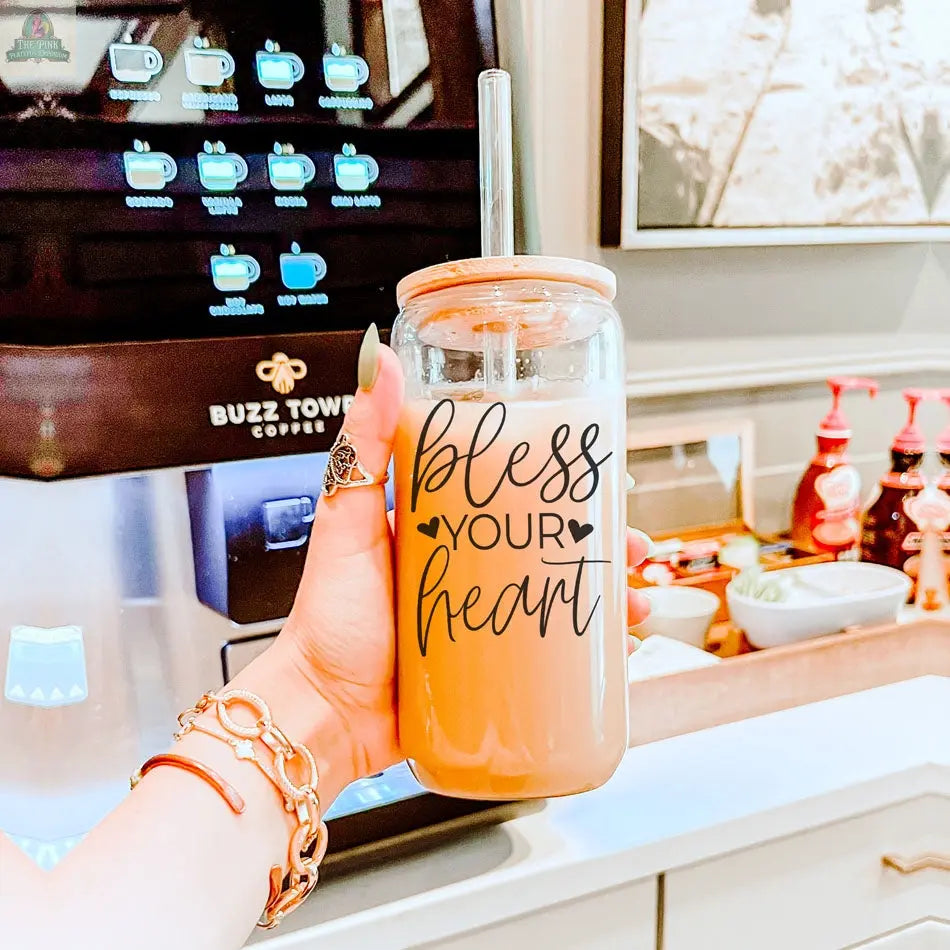 A hand holds the Bless Your Heart 20oz glass cup filled with iced coffee and a straw, in front of a coffee machine and syrup bottles on the counter.