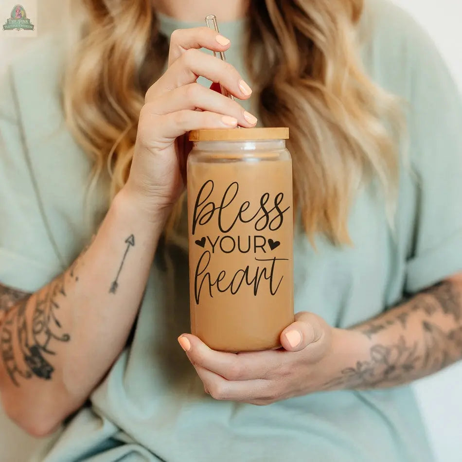 A woman with wavy blonde hair and tattooed arms holds a Bless Your Heart 20oz glass cup—an iced coffee in a glass jar with a bamboo lid and straw, featuring script and heart accents.