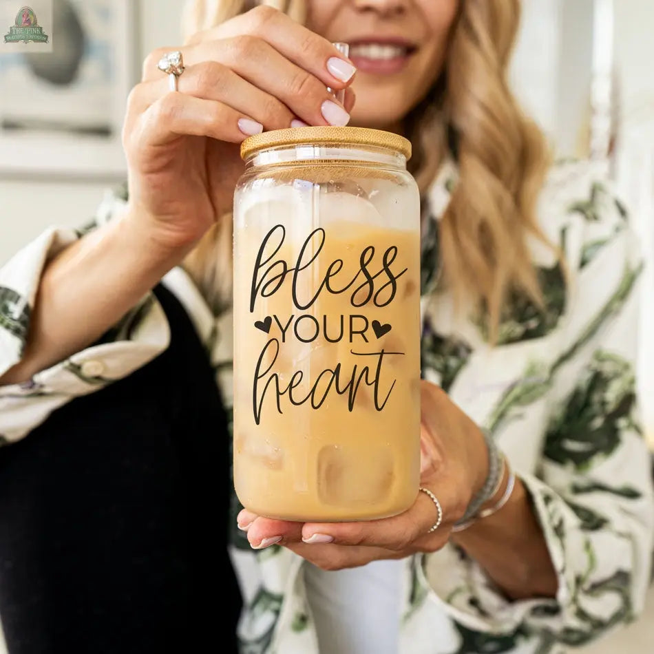 A woman in a patterned shirt holds the Bless Your Heart 20oz glass cup with a bamboo lid, filled with iced coffee. Her hand, drink, and part of her smiling face are visible, showing off the playful message and charming heart graphics.
