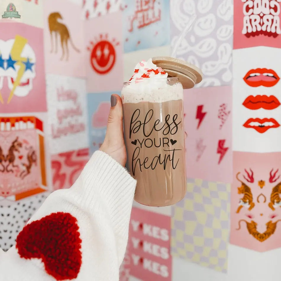 A hand in a white sweater with red hearts holds a Bless Your Heart 20oz glass cup of iced coffee topped with whipped cream. The jar displays playful lettering, while colorful art prints liven up the background wall.