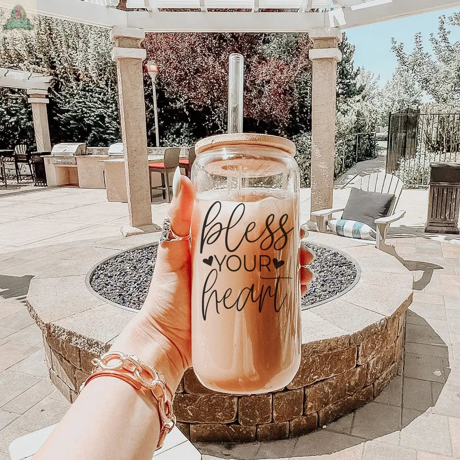 A hand holds the Bless Your Heart 20oz glass cup with a bamboo lid and straw in front of a fire pit and patio seating on a sunny patio.