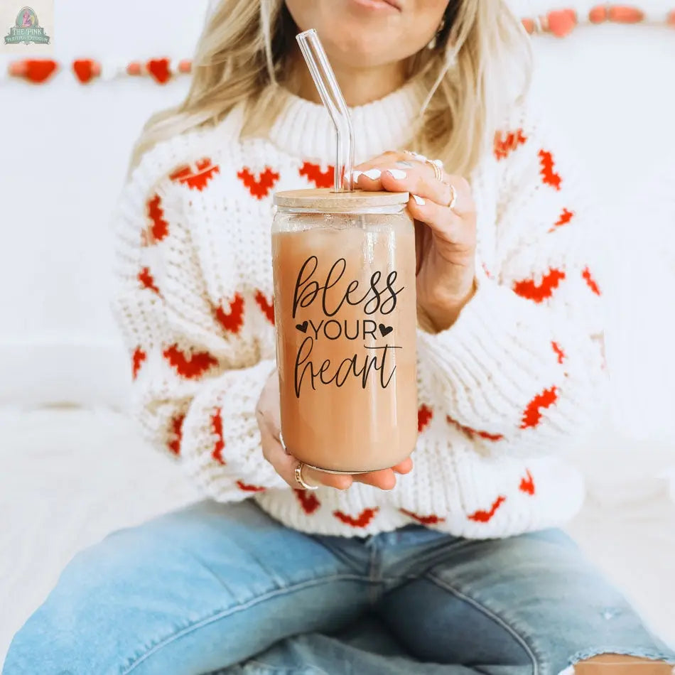 Someone in a white sweater with red hearts holds a Bless Your Heart 20oz glass cup filled with iced coffee and a clear straw, sitting cross-legged on a bed or couch.