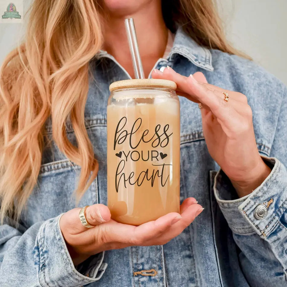 A woman in a denim jacket holds the Bless Your Heart 20oz glass cup with a bamboo lid and metal straw. The mason jar, filled with a light brown drink, showcases decorative lettering on the front.