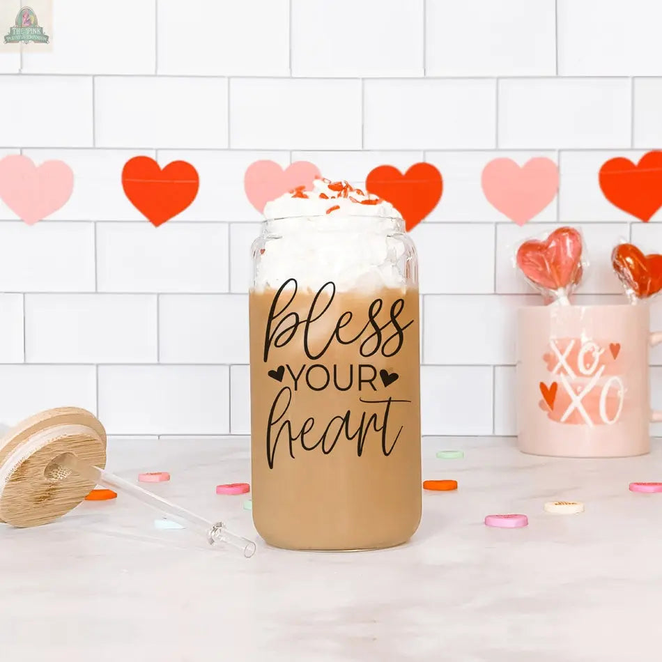 A Bless Your Heart 20oz glass cup filled with iced coffee, whipped cream, and heart-shaped sprinkles rests on a white counter accented with heart decor and a lollipop cup in the background.