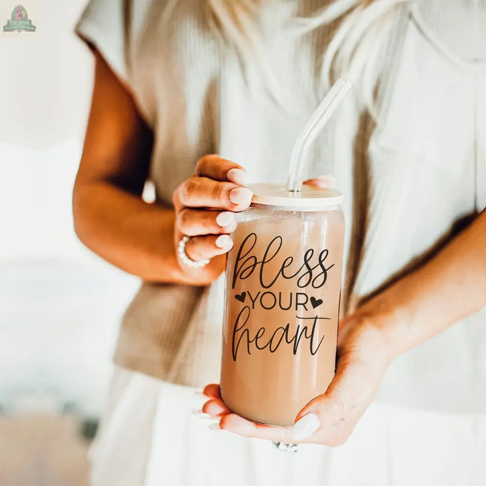 A woman with long blonde hair holds the Bless Your Heart 20oz glass cup filled with iced coffee or tea, featuring a reusable straw and "bless your heart" in script on the jar.