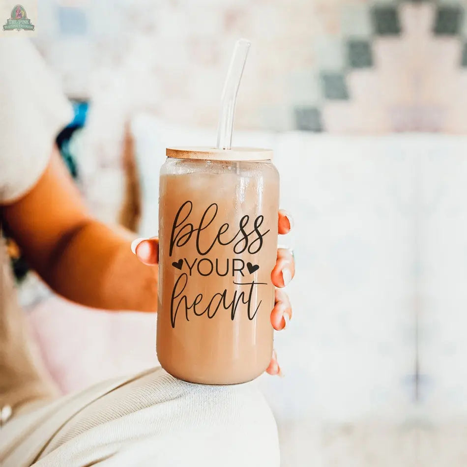 A person holds the Bless Your Heart 20oz glass cup with a clear straw and bamboo lid, filled with iced coffee or tea. The cup features "Bless Your Heart" in black cursive and small heart designs.