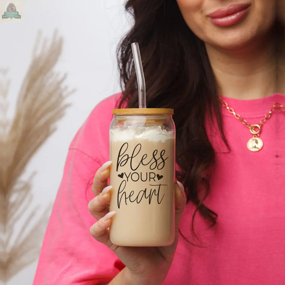 A woman in a pink shirt holds the Bless Your Heart 20oz glass cup with a decorative bamboo lid and straw, adding charm. She pairs it with a gold necklace featuring a round pendant.