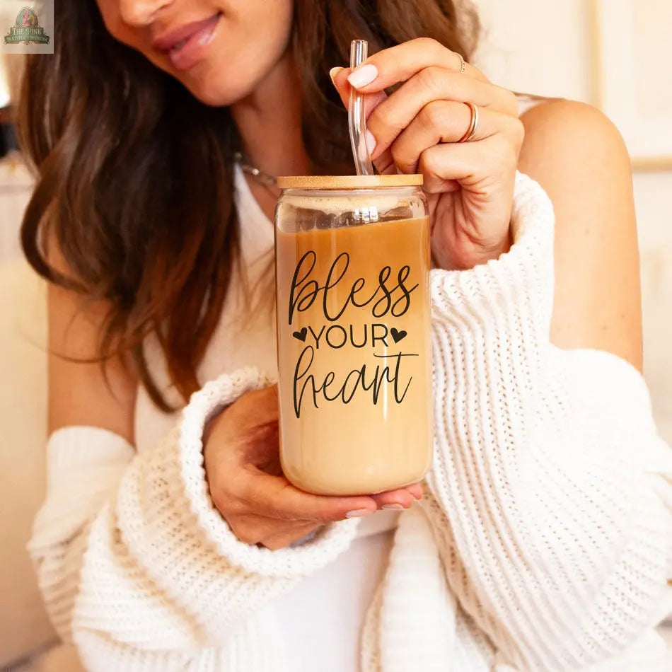 A woman in a white sweater holds an iced coffee in a Bless Your Heart 20oz glass cup with a straw, its decorative script adding charm to her drink.