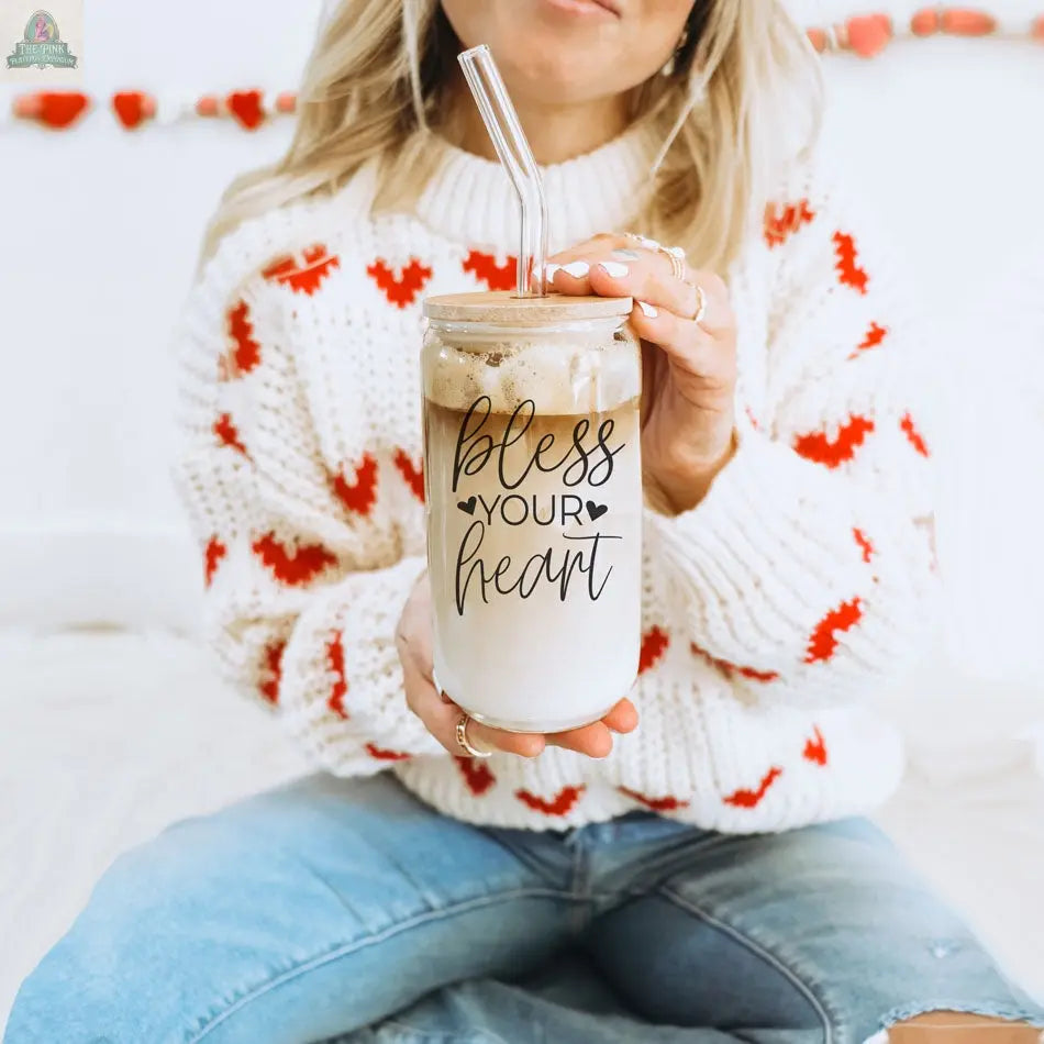 A woman in blue jeans and a white sweater with red hearts sits on a bed, holding the Bless Your Heart 20oz glass cup filled with iced coffee, with a festive red heart garland in the background.
