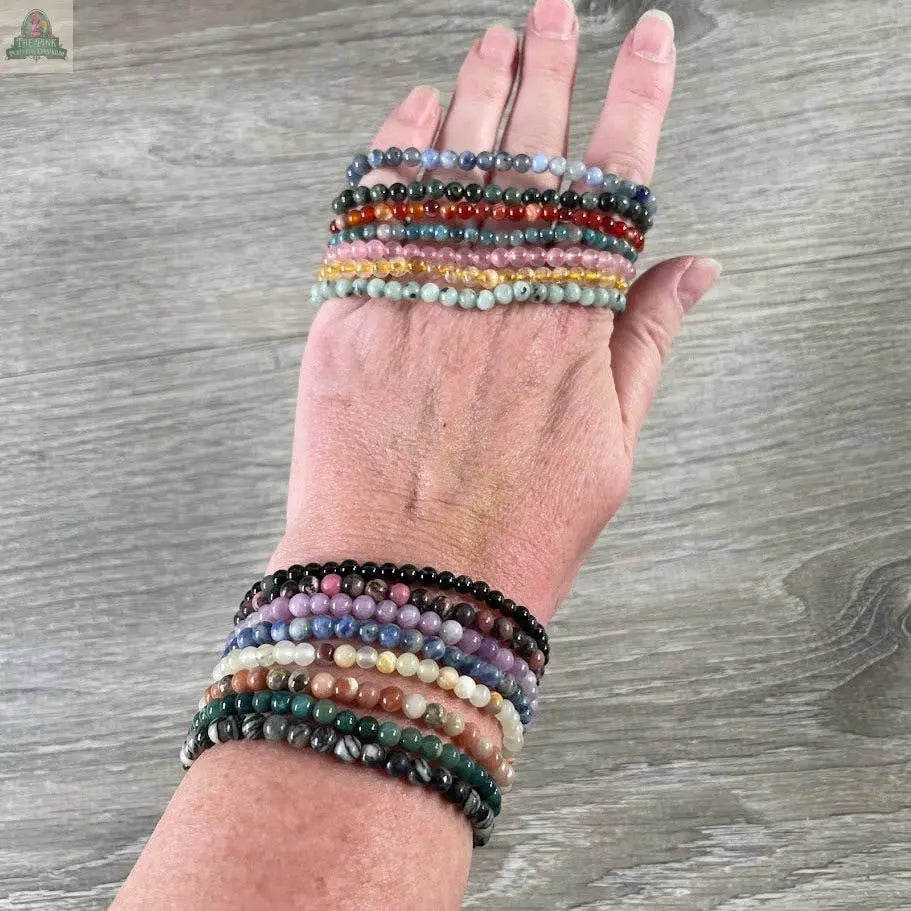 A hand displays vibrant Keystone Crystals gemstone bead bracelets (4mm) from Pink Platypus Emporium—genuine healing crystal jewelry in various colors over a wooden surface.