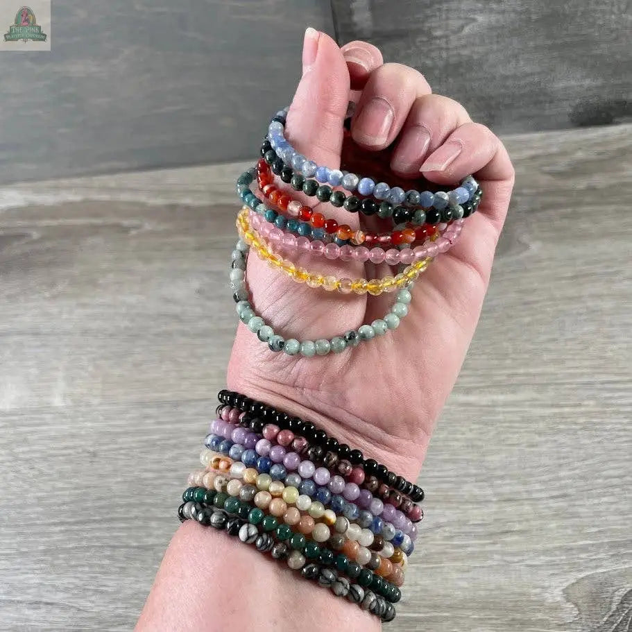 A hand wears several Keystone Crystals gemstone bead bracelet 4mm– Genuine Healing Crystal Jewelry from Pink Platypus Emporium, holding more beaded bracelets against a wooden surface background.