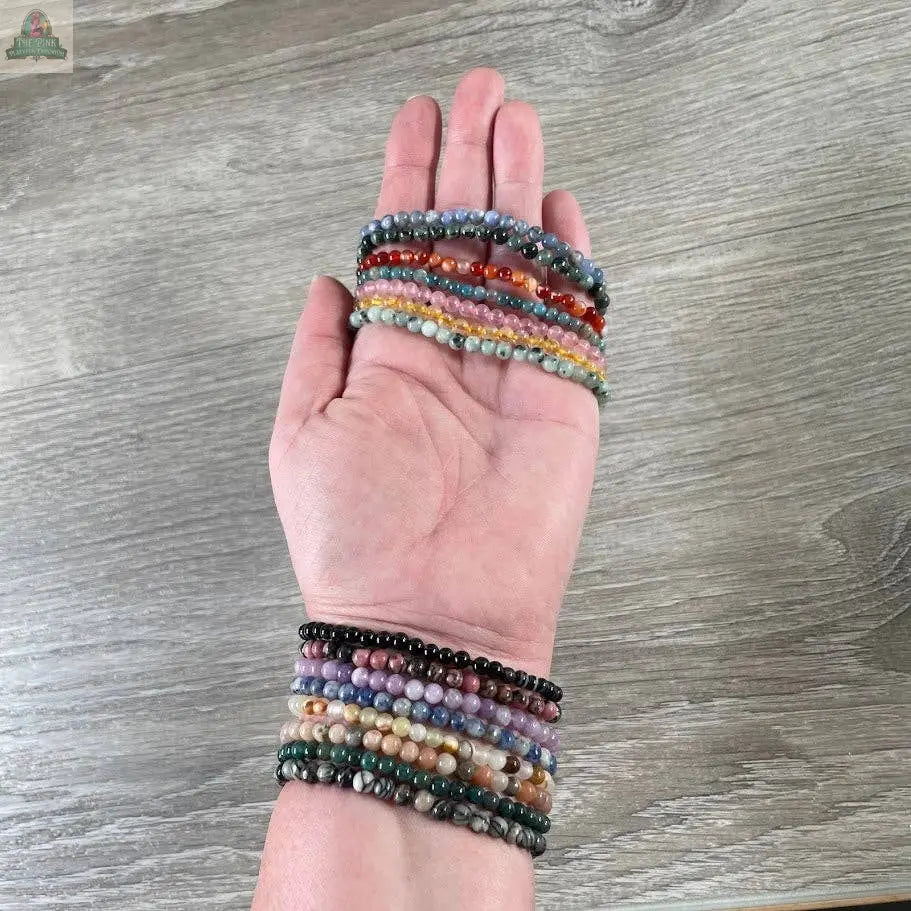 A hand showcases Keystone Crystals’ 4mm gemstone bead bracelet—genuine healing crystal jewelry from Pink Platypus Emporium—stacked stylishly against a wood-textured background.