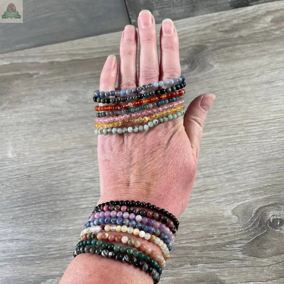 A hand displays Keystone Crystals' 4mm gemstone bead bracelets from Pink Platypus Emporium, featuring vibrant genuine healing crystals in green, red, purple, yellow, and black against a wooden background.