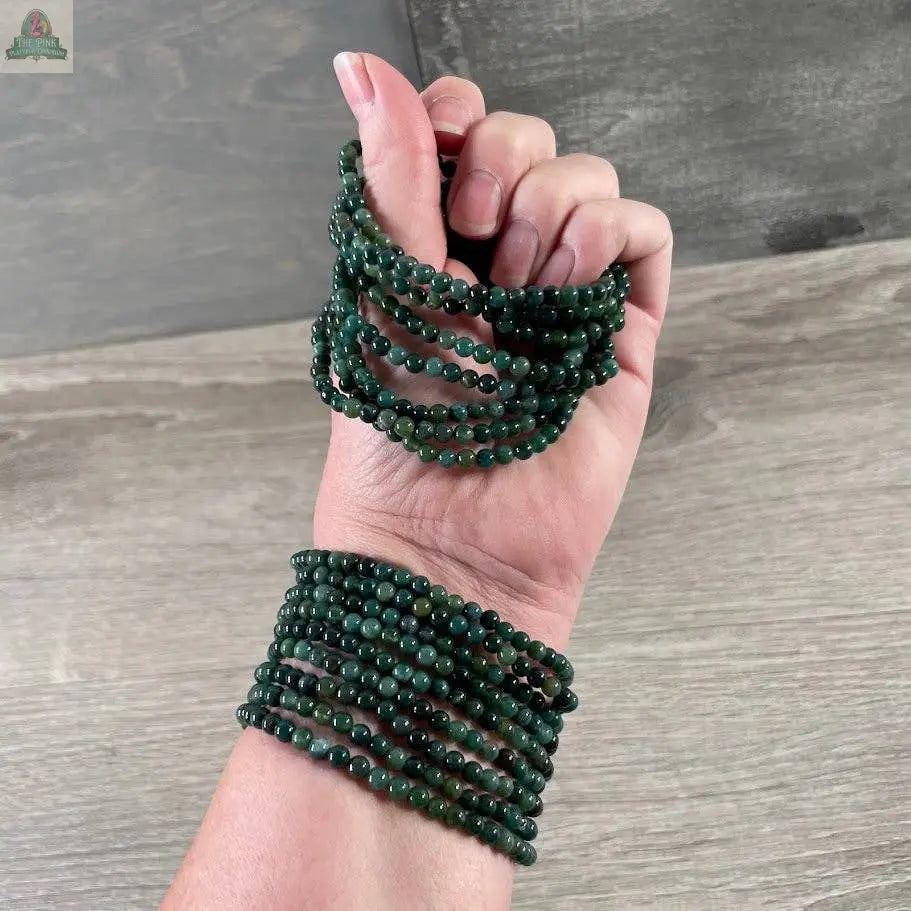 A hand and wrist wearing Keystone Crystals’ 4mm gemstone bead bracelet—Genuine Healing Crystal Jewelry from Pink Platypus Emporium—are shown against a wood-textured background.