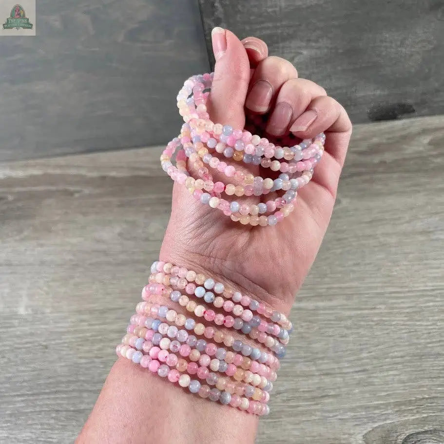 A hand wearing and holding multiple Keystone Crystals 4mm gemstone bead bracelets—Genuine Healing Crystal Jewelry from Pink Platypus Emporium—displayed against a gray and wooden background.
