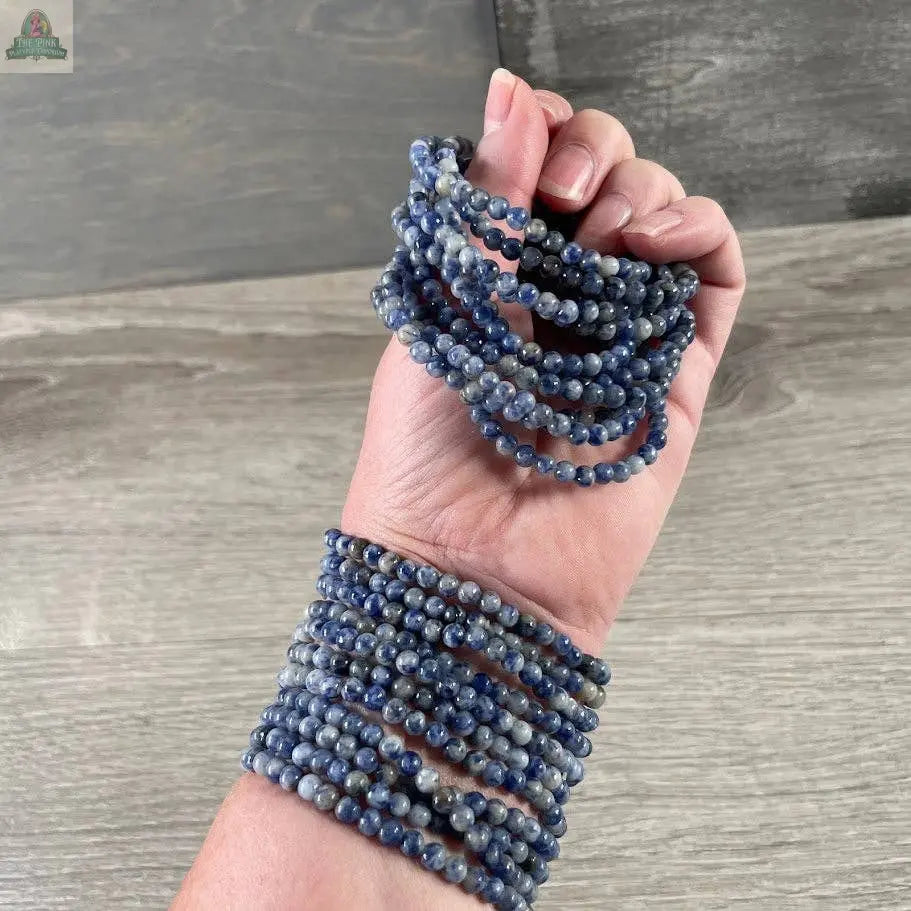 A hand and wrist adorned with Keystone Crystals’ 4mm gemstone bead bracelet from Pink Platypus Emporium, featuring genuine healing crystals, are set against a wooden background to highlight this stylish jewelry design.