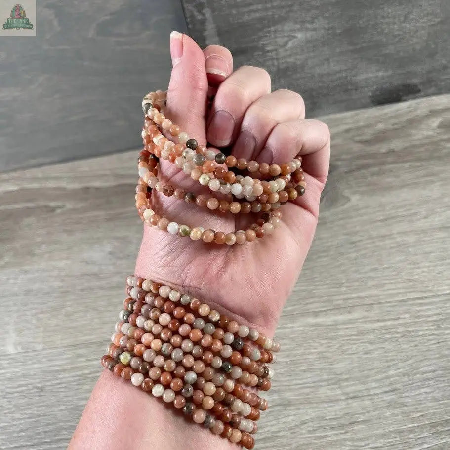 A hand wears several strands of Keystone Crystals’ 4mm gemstone bead bracelet—Genuine Healing Crystal Jewelry from Pink Platypus Emporium—showcasing round, polished beads against a wooden background.