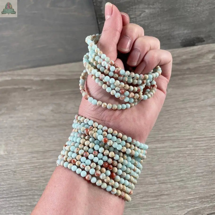 A hand displays Keystone Crystals’ gemstone bead bracelet 4mm—Genuine Healing Crystal Jewelry from Pink Platypus Emporium—featuring round, polished amazonite beads in light blue, cream, and brown against a wooden background.