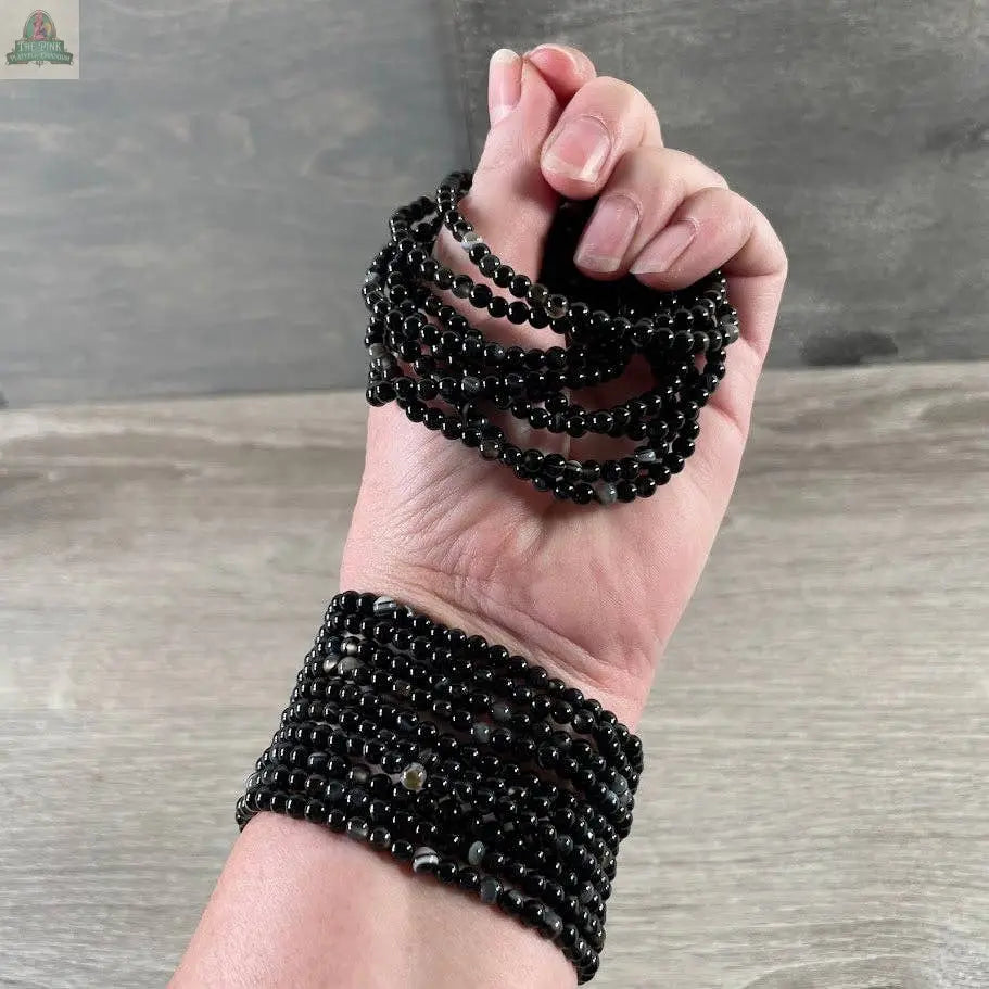 A light-skinned hand showcases Keystone Crystals’ 4mm gemstone bead bracelet—Genuine Healing Crystal Jewelry from Pink Platypus Emporium—worn in shiny black strands for a layered look against a gray background.