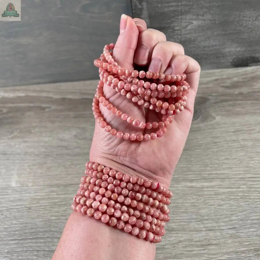 A hand and wrist adorned with several Keystone Crystals gemstone bead bracelet 4mm–Genuine Healing Crystal Jewelry in pink from Pink Platypus Emporium, their marbled beads coiling elegantly against a wooden background.