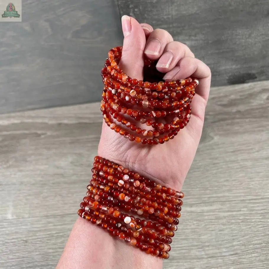 A hand wears multiple stacked Keystone Crystals gemstone bead bracelets (4mm) from Pink Platypus Emporium, featuring round, polished healing crystal beads. More strands drape over the fingers, resting on a wooden surface in the background.