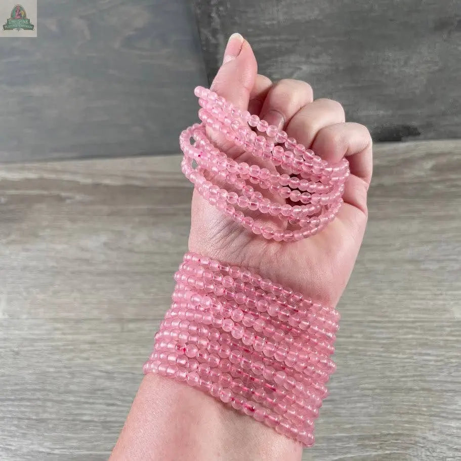 A hand and wrist are adorned with multiple Keystone Crystals 4mm gemstone bead bracelets—Genuine Healing Crystal Jewelry from Pink Platypus Emporium—featuring small, round pink beads against a wooden surface.