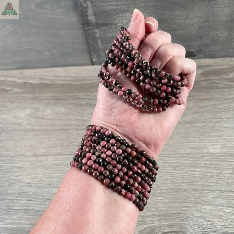 A hand adorned with Keystone Crystals' 4mm gemstone bead bracelets from Pink Platypus Emporium, in pink, red, and black hues, is displayed on wood—highlighting the allure of genuine healing crystal jewelry.
