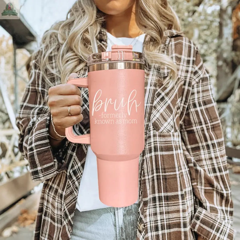 A woman in a plaid shirt holds a Bruh Pink 40oz tumbler with handle, the perfect mom gift. The large pink cup stands out as she relaxes outdoors by the trees and a wooden bench.
