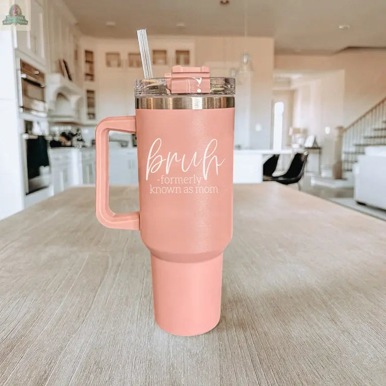 The Bruh Pink 40oz tumbler with handle sits on a wooden table in a modern kitchen. It features “bruh - formerly known as mom” in white text, a clear lid with straw, and makes a perfect mom gift for any occasion.