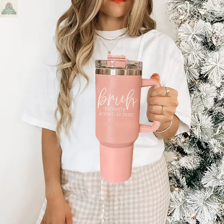 A woman with long wavy hair in a white shirt and checkered pants holds the Bruh Pink 40oz tumbler with handle, reading "Bruh Formerly Known as Mom," beside a snow-dusted Christmas tree—the perfect holiday gift for moms.