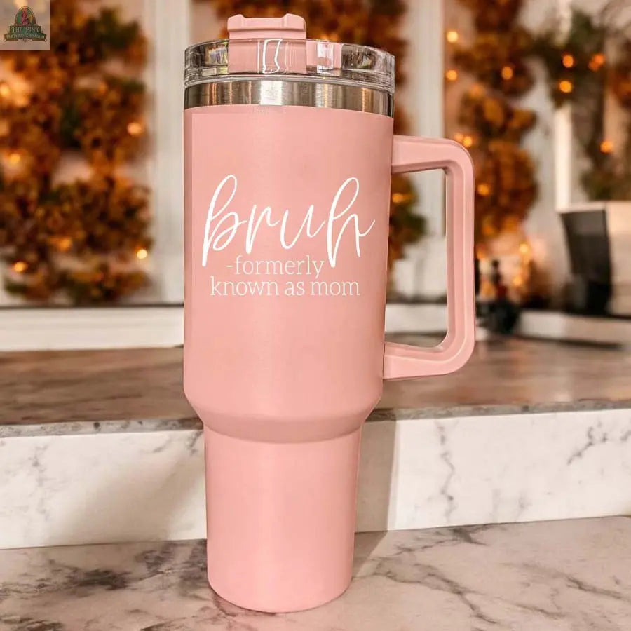 A Bruh Pink 40oz tumbler with a handle rests on a marble counter. White text reads "Bruh Formerly Known as Mom"—a perfect gift for moms. The warmly blurred, soft decor creates a cozy vibe.