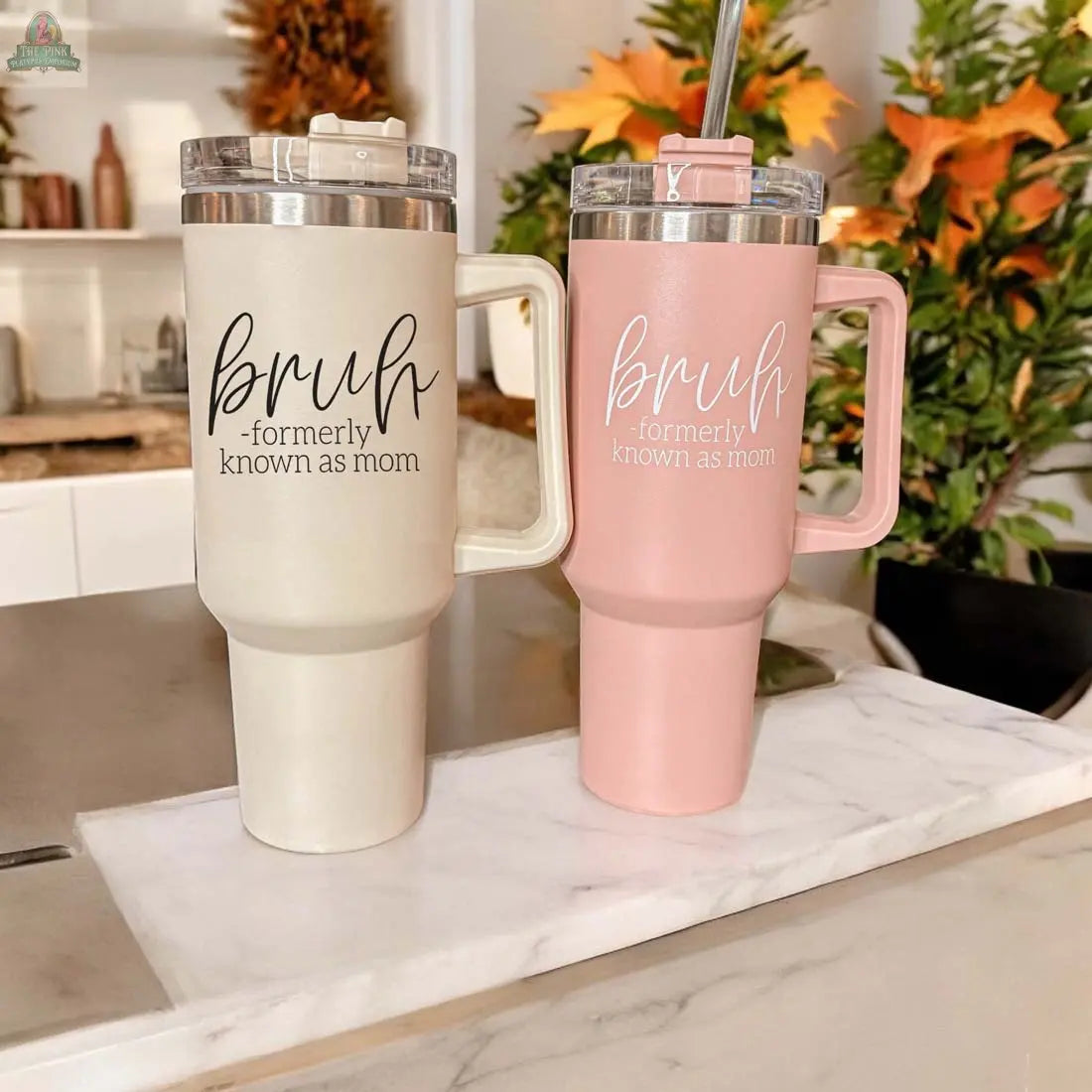 The Bruh Pink 40oz tumbler with handle sits on a marble tray, featuring “bruh - formerly known as mom”—a perfect mom gift. Fall-themed decor and kitchen items accent the background.