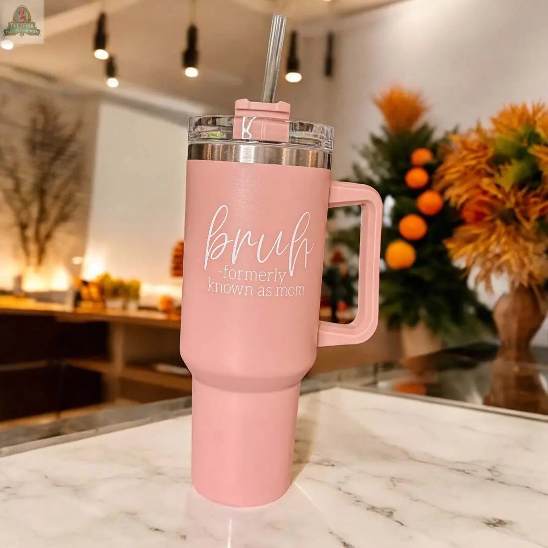 The Bruh Pink 40oz tumbler with handle sits on a marble countertop, displaying "bruh - formerly known as mom" in white text. Ideal as a mom gift, the cozy scene features plants and orange accents in the background.