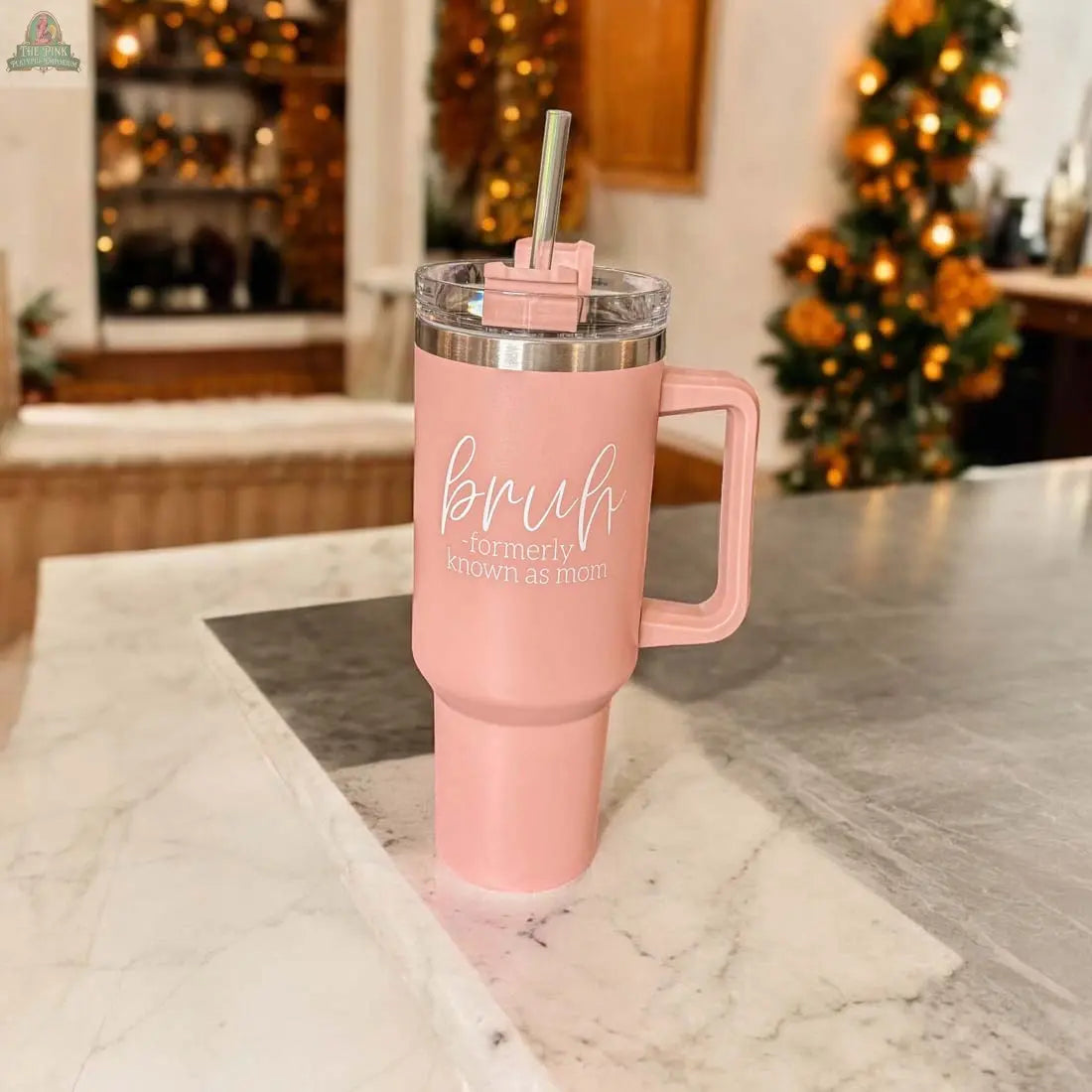 The Bruh Pink 40oz tumbler with handle and straw, printed with "Bruh Formerly Known as Mom," sits on a marble countertop—an ideal mom gift. Holiday lights and blurred decorations create a festive background.