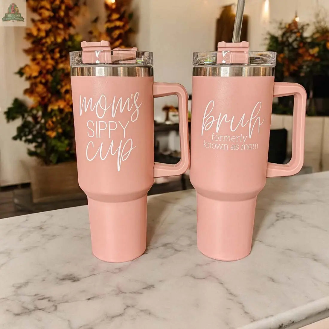 Two pink travel mugs, including the Bruh Pink 40oz tumbler with handle—ideal as a mom gift—sit on a marble counter, set against a backdrop of warm, blurred autumn foliage.