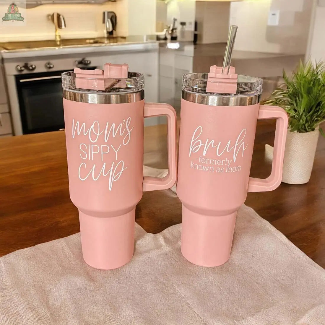 Two Bruh Pink 40oz tumblers with handles sit on a kitchen table—one says “moms sippy cup,” the other “Bruh Formerly Known as Mom.” Perfect mom gift, with kitchen and plant in background.