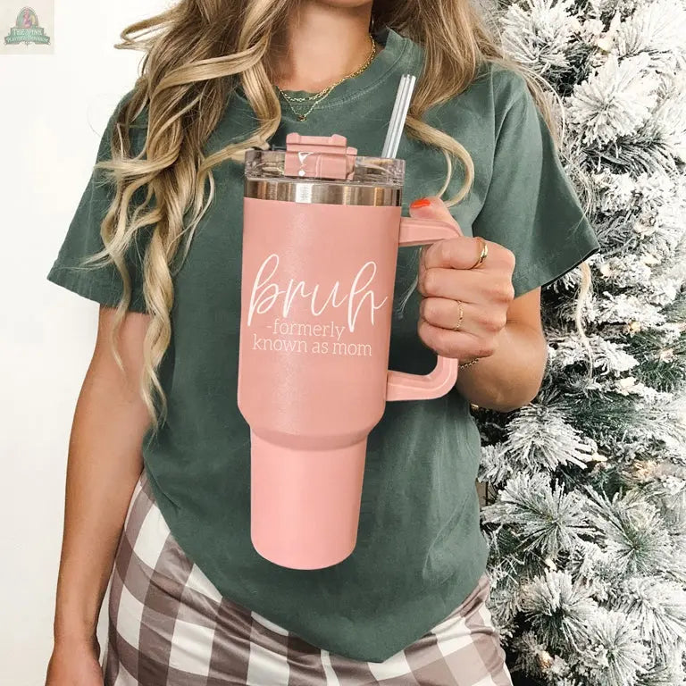 A woman in a green shirt and plaid pants holds the Bruh Pink 40oz tumbler with handle reading "Bruh Formerly Known as Mom." A snowy Christmas tree in the background makes it a perfect holiday gift for moms.
